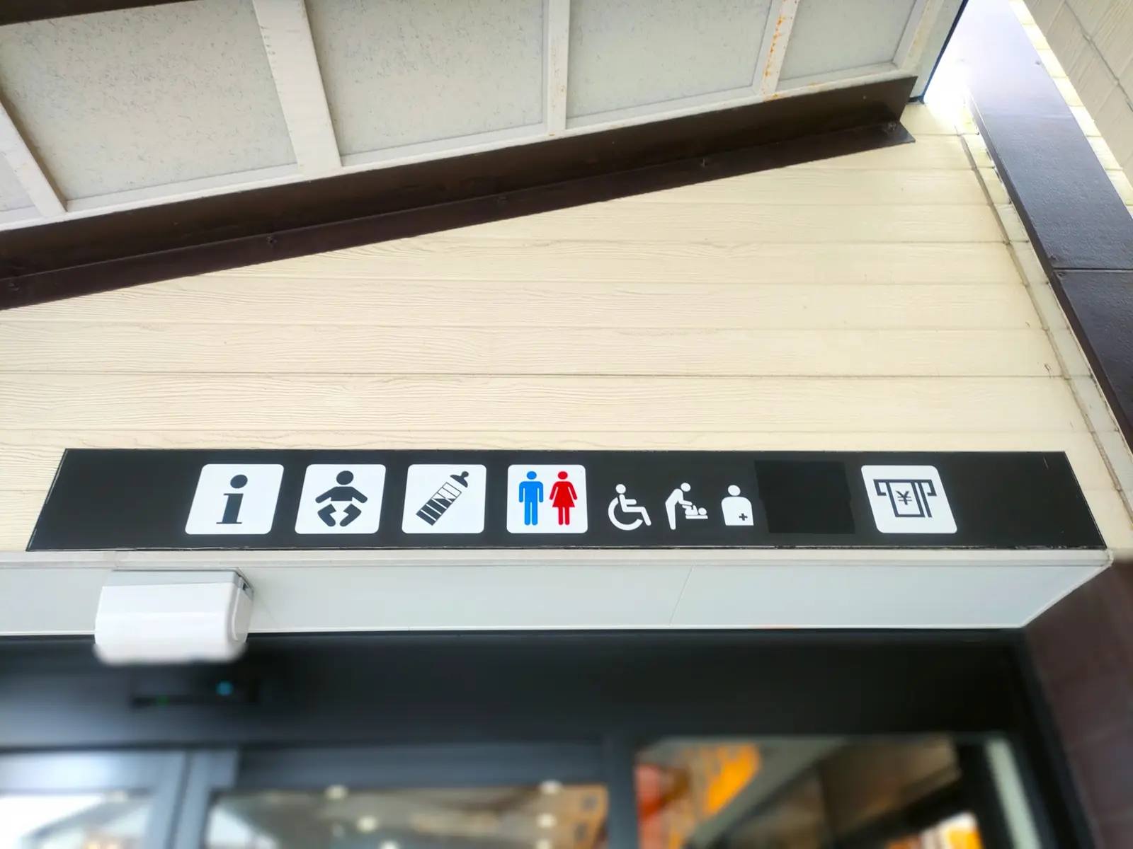 Ein Hinweisschild über einem Eingang in Japan zeigt mit Piktogrammen die verfügbaren barrierefreien Einrichtungen an – darunter Rollstuhl-Zugang, Mehrzwecktoilette, Wickelplatz und Erste Hilfe.