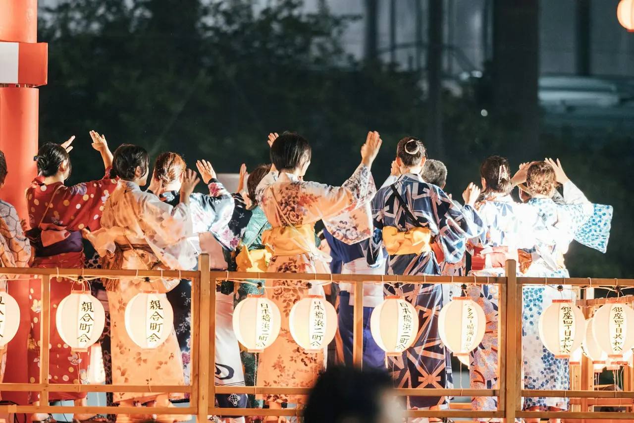 Dansere i fargerike yukata løfter hendene på en treplattform, hvite papirlykter henger på rekkverket.