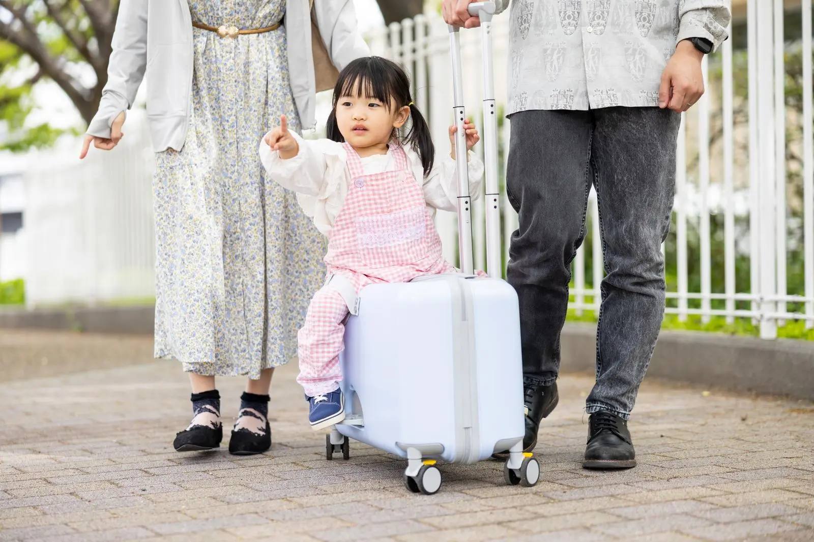 Ein kleines Mädchen sitzt auf einem Rollkoffer zwischen seinen Eltern und zeigt nach vorne, unterwegs auf einem Weg in Japan – Sinnbild für eine Familienreise mit Kindern.