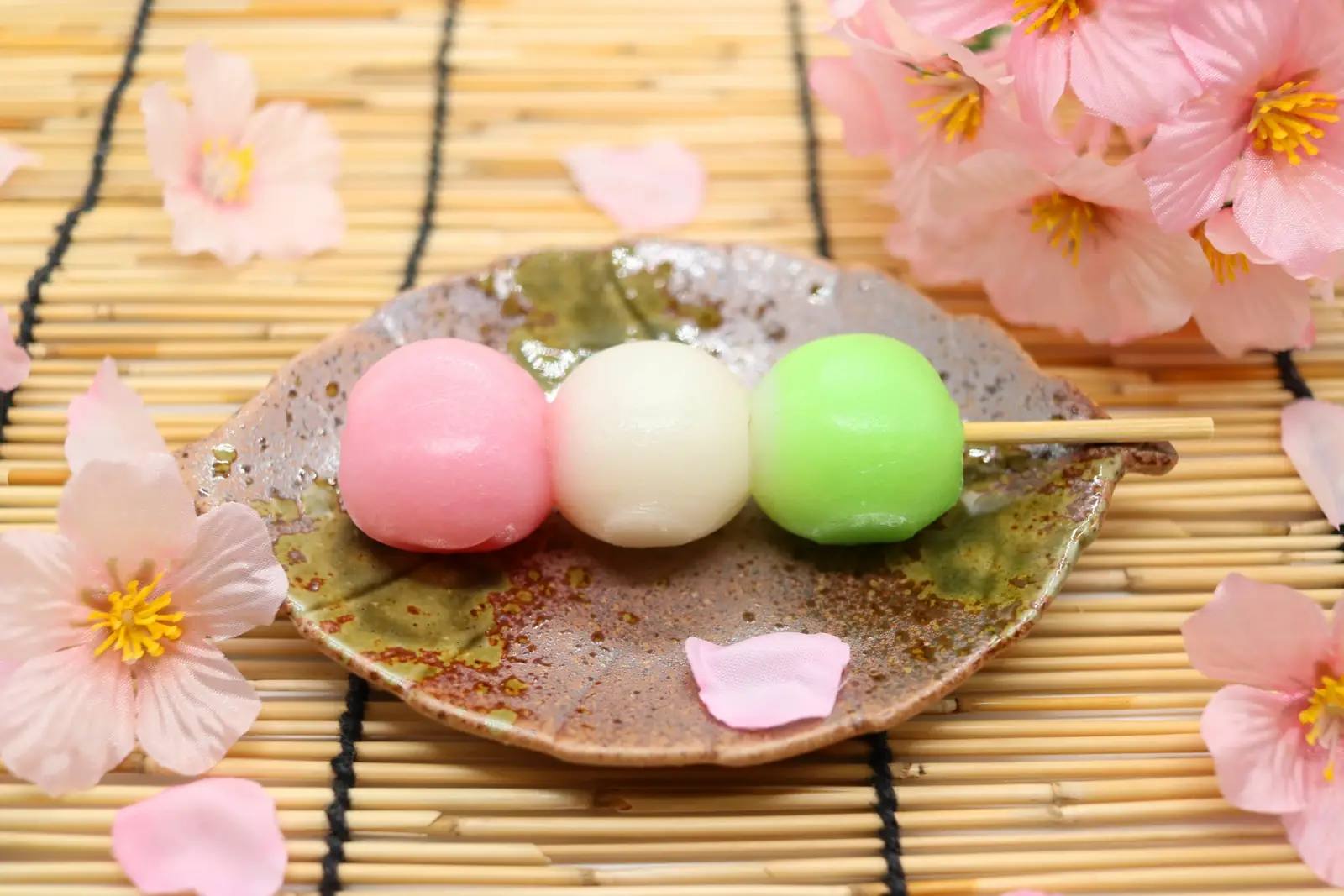 Hanami-dango – drei Reisklöße in Rosa, Weiß und Grün auf einem Spieß, daneben Kirschblüten