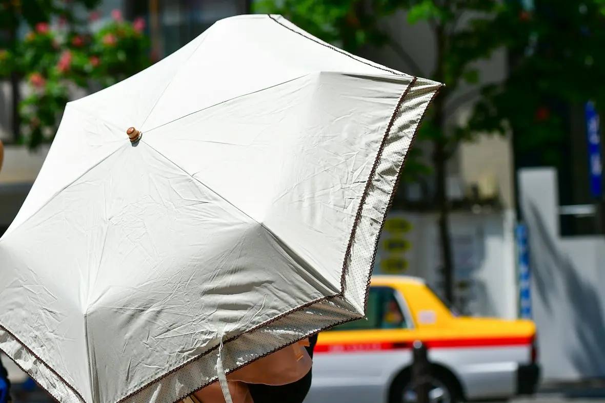Iemand loopt met een lichte parasol over een zonnig trottoir in een Japanse stad; op de achtergrond rijdt een geel-rode taxi.