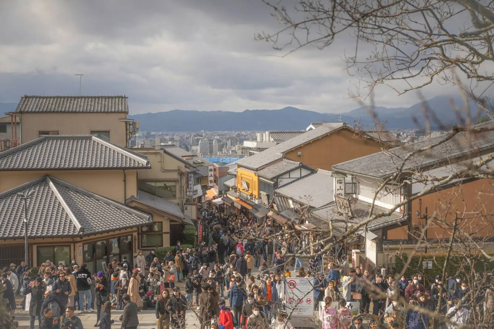 Blik over de daken van de oude stad aan de opgang naar de Kiyomizu-dera in Kyoto op een bewolkte winterdag; in de steeg verdringen zich mensen in winterjassen en donsjassen, sneeuw ligt er niet.