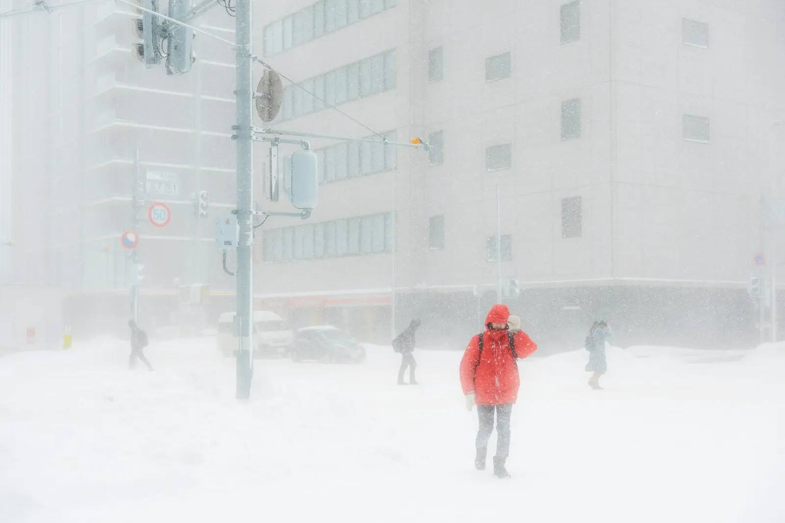 Sneeuwstorm op een kruispunt in Sapporo: iemand in een rode winterjas met capuchon en handschoenen ploetert door diepe sneeuw, op de achtergrond worstelen andere voetgangers zich vooruit.