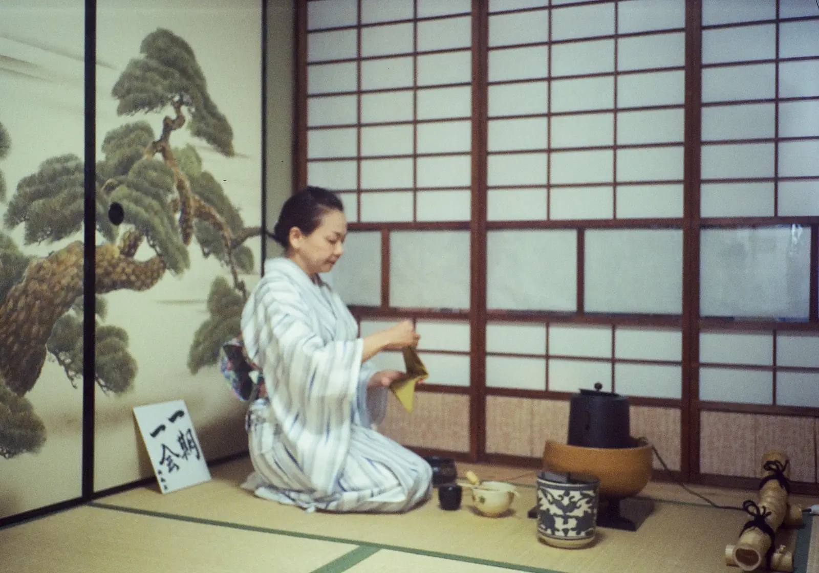 Eine Teemeisterin im Kimono bereitet in einem traditionellen Teeraum Matcha zu; im Hintergrund die Kalligrafie ichigo ichie