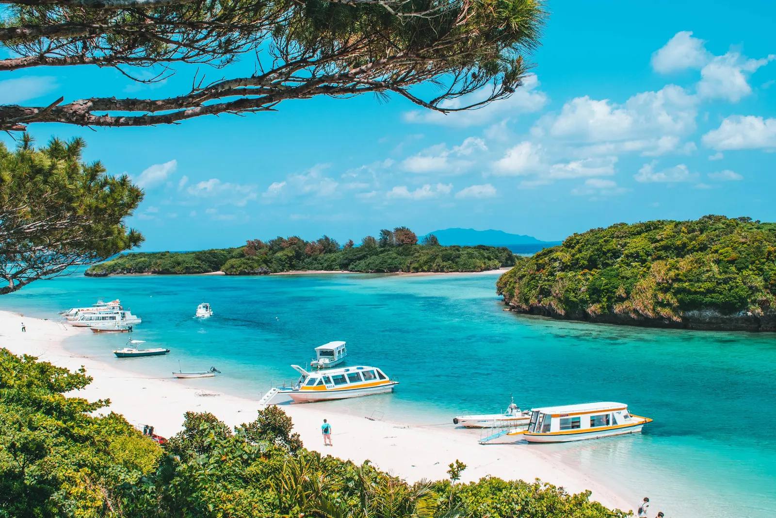 Glasbodenboote liegen am weißen Sandstrand der Kabira-Bucht auf Ishigaki, dahinter türkisfarbenes Wasser und dicht bewaldete Inseln
