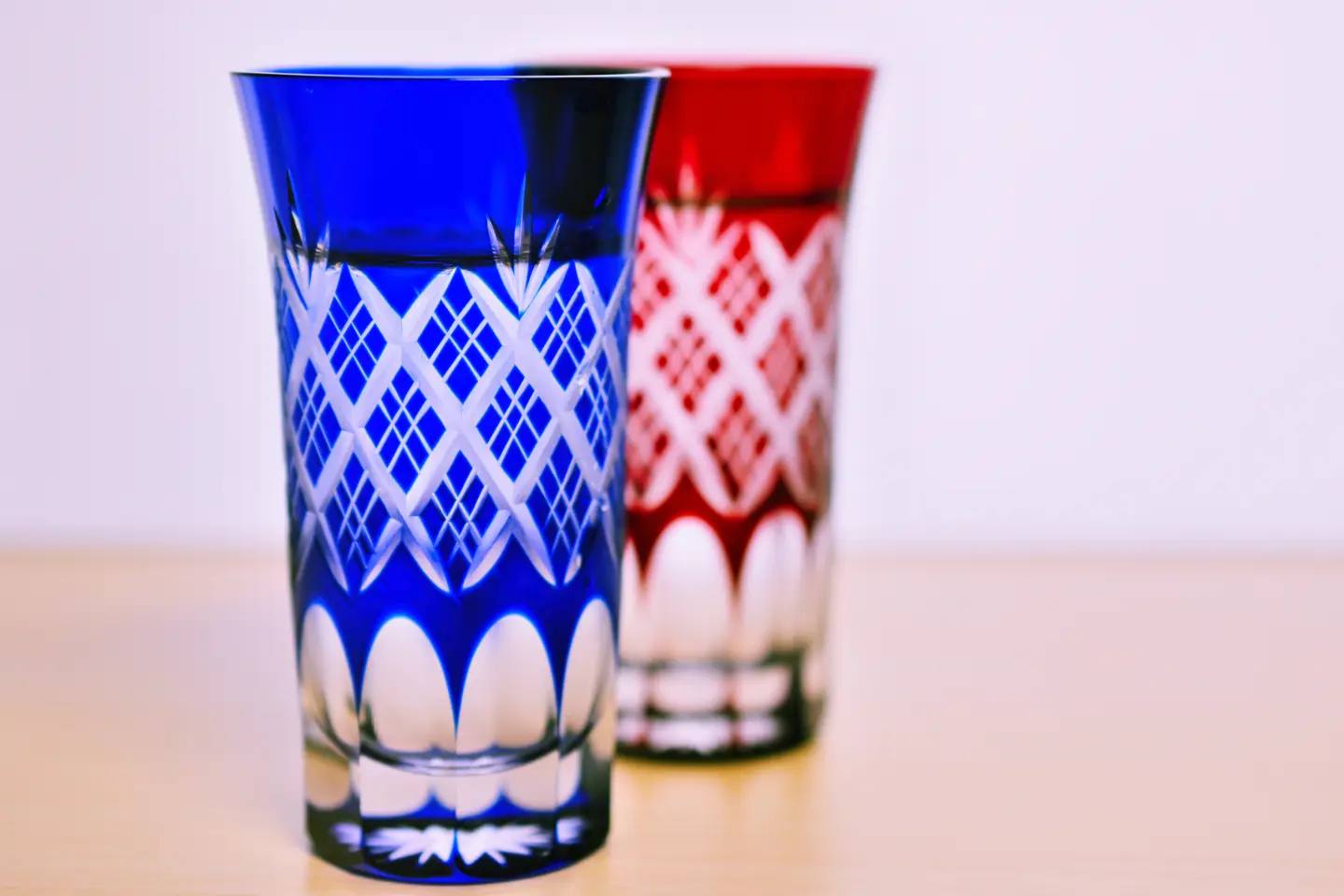 Two Edo-Kiriko glasses in blue and red with cut diamond and fan patterns, standing on a light wooden table.