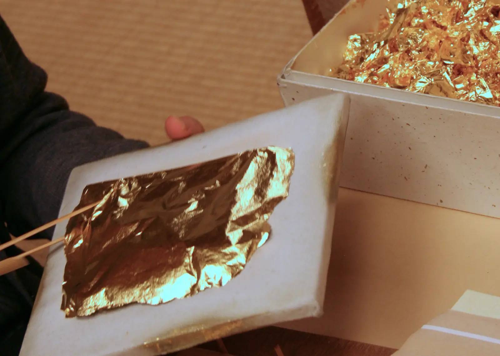 Wafer-thin gold leaf lies on a sheet of paper and is picked up with two bamboo sticks; beside it stands a bowl with more sheets of gold, in a workshop in Kanazawa.