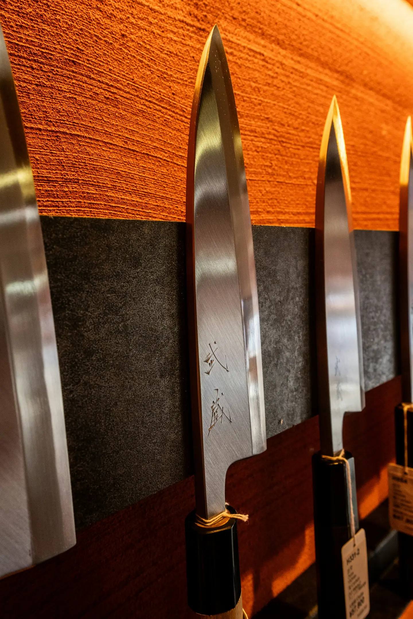 Several Japanese kitchen knives hanging side by side on a shop wall; characters are engraved in the blade of the front knife.