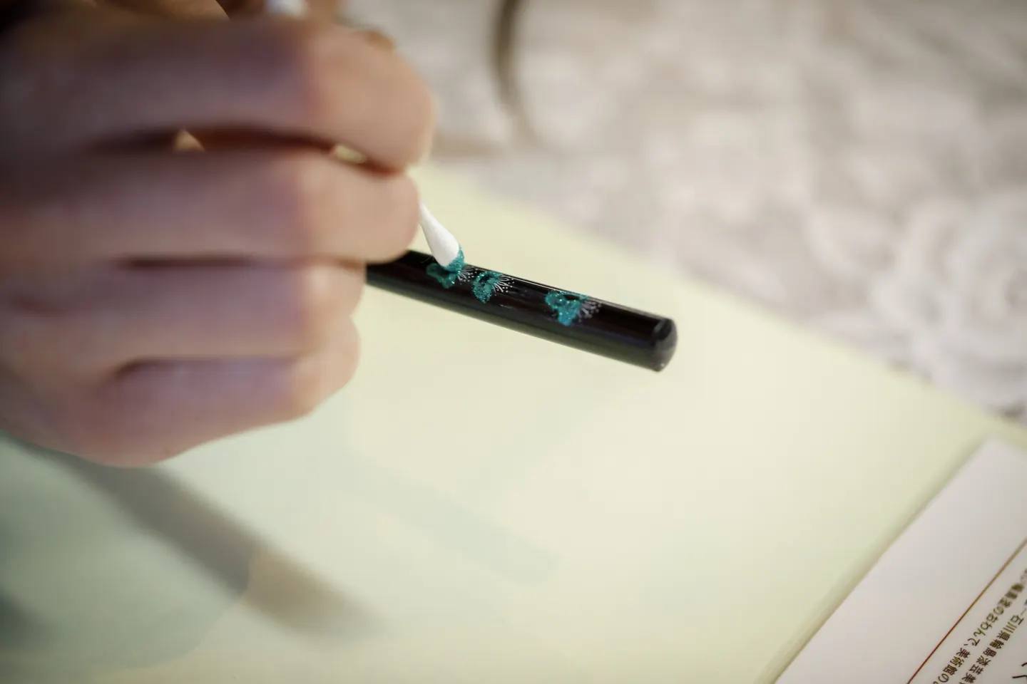 A hand applying turquoise powder to a black-lacquered chopstick with a cotton bud; the blossom pattern emerges as it does so.