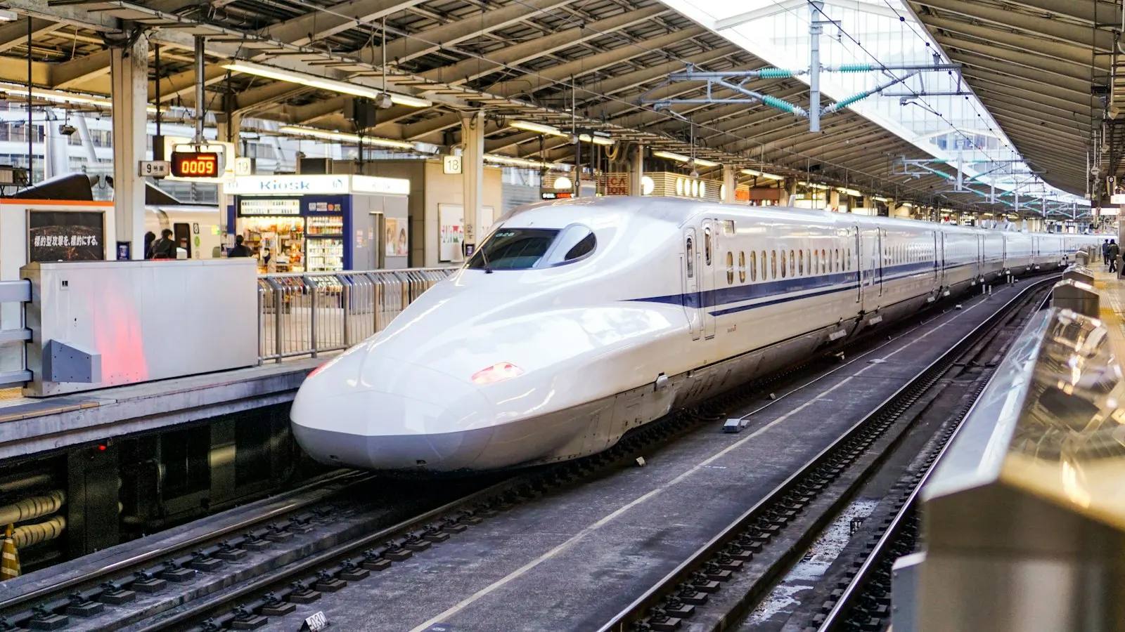 Ein weißer Shinkansen mit blauem Streifen steht am überdachten Bahnsteig im Bahnhof Tokio, links ein Kiosk und Bahnsteiganzeigen.