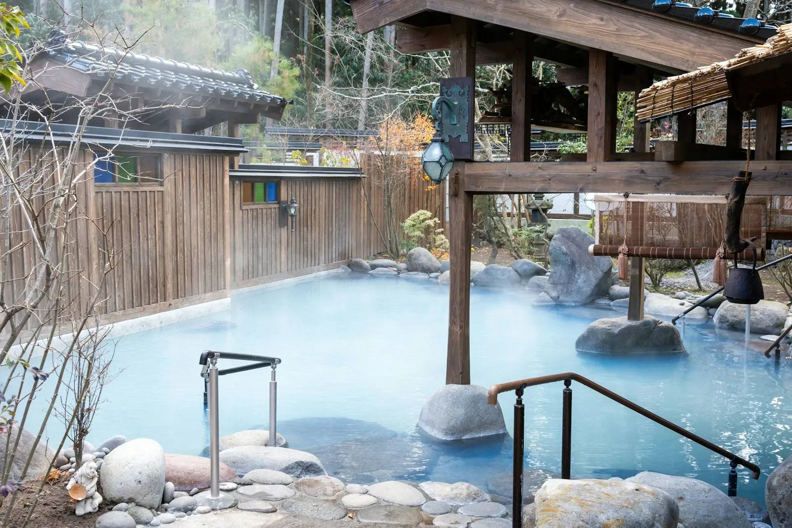 Dampfendes Freiluftbad aus Natursteinen mit milchig-blauem Wasser, überdacht von einem Holzpavillon und umgeben von Holzzäunen und Bäumen.