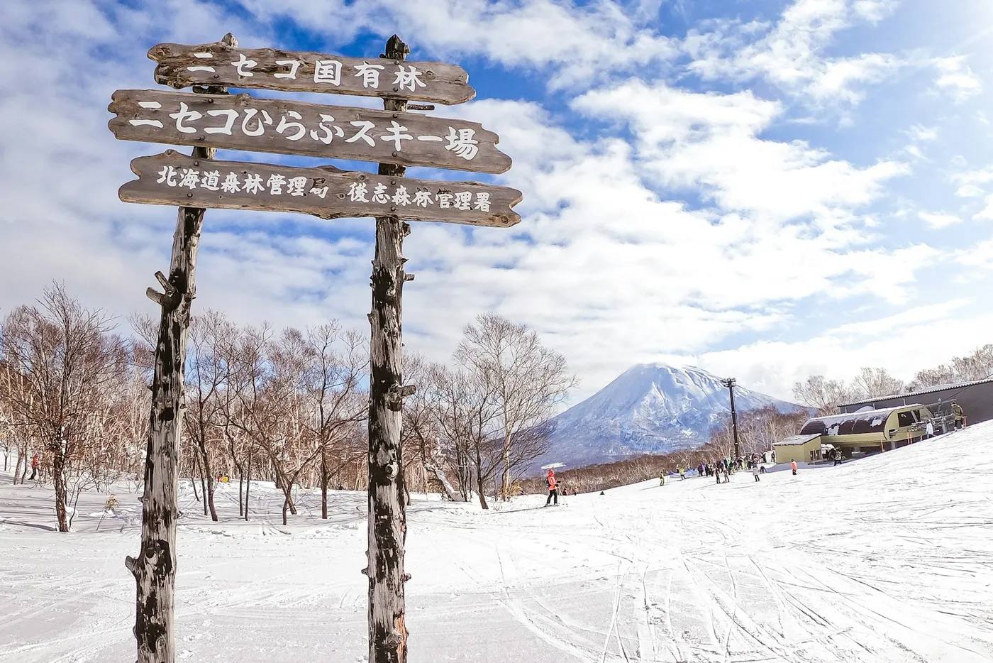 Holzschild des Skigebiets Niseko Hirafu im Schnee, dahinter der markante Vulkankegel des Berges Yōtei und die Piste bei blauem Himmel auf Hokkaidō